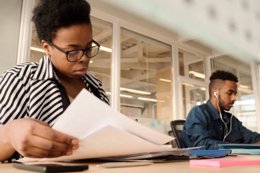 Modern ofiste kağıtları düzenleyen şık bir Afrikalı Amerikalı kadın. Bu arada genç erkek arkadaşı bilgisayar üzerinde çalışıyor. Düşük açılı.