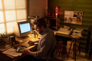 Masada çalışan beyaz bir adam klasik bilgisayar ve hesap makinesi kullanıyor. Evrak işleri ve ofis malzemeleriyle çevrili.