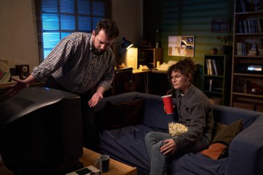 Beyaz adam ayakta dikilip televizyona doğru el kol hareketi yaparken beyaz kadın elinde bir kase patlamış mısırla kanepede oturup kırık ekran seyrediyor.