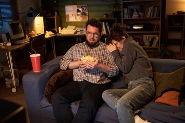 Kafkasyalı bir adam elinde patlamış mısırla koltukta oturmuş, omuzlarına yaslanmış beyaz saçlı bir kadının olduğu korku filmi seyrediyor.