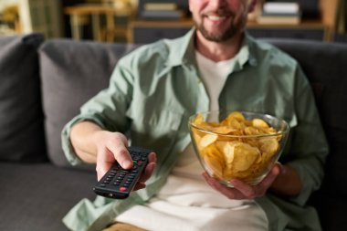 Kafkasyalı genç adam koltukta oturmuş elinde cam patates cipsi tutarak uzaktan kumandayla gülümsüyor ve oturma odasında televizyon seyrediyor.