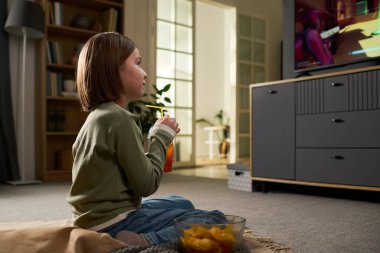 Kafkasyalı çocuk televizyonda çizgi film seyrederken samandan meyve suyu içiyor, yakınlarda bir kase abur cubur var ve oturma odasındaki ekrana odaklanıyor.