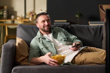 Kafkasyalı adam, elinde uzaktan kumanda ve patates cipsi tutarak kanepeye yaslanıyor ve modern oturma odasında rahatça gülümseyen televizyon seyrediyor.