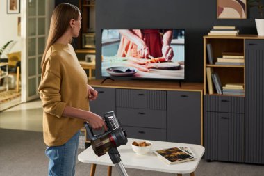 Kafkasyalı genç bir kadın elinde kablosuz süpürgeyle televizyon ekranına bakarken modern oturma odasındaki sebzeleri doğrayan ellerin yakın çekimini gösteriyor.