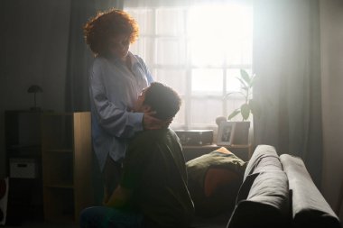 Orta yaşlı, beyaz bir kadın koltukta oturan beyaz bir gencin yüzünü nazikçe tutuyor. İkisi de oturma odasında sevgi dolu bir anı paylaşıyor.