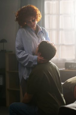 Kafkasyalı genç bir kadın ayakta duruyor ve beyaz bir gencin yüzüne nazikçe dokunuyor. Diz çöküyor ve arka planda penceresi olan yatak odasında hafifçe ona bakıyor.