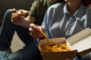 Birlikte oturan genç çift pizza ve patates kızartması yiyor, fast food yemeğini paylaşıyor, eller ve kısmi yüzler görünür, ev ortamında gelişigüzel takılıyor.