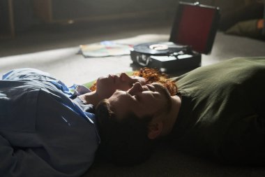 Çift yerde gözleri kapalı yatıyordu vinil plak çaların yanında, yumuşak ışıkta birlikte dinleniyorlardı, evde huzurlu bir anı paylaşıyorlardı.