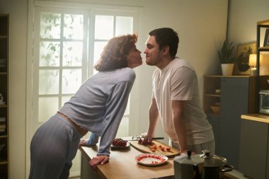 Beyaz, genç, yetişkin bir kadın mutfakta gülümseyen ve göz teması kuran beyaz bir adamı öpmek için öne eğiliyor.