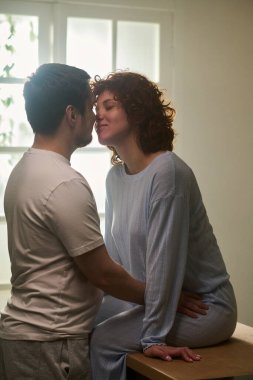 Masada oturan kadını kucaklayan, gözleri kapalı gülümseyen, kadının beline nazikçe dokunan, aydınlık odada samimi bir anın dikey görüntüsü.