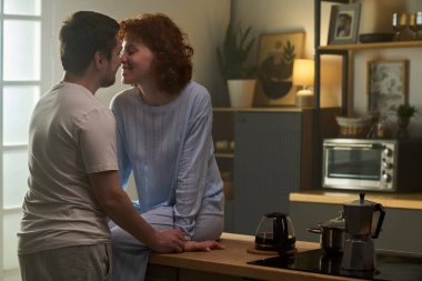 Kafkasyalı genç bir adam mutfak tezgahında oturan beyaz bir kadının yanında duruyor. Gülümsüyor ve öpüşmek için eğiliyor. Sabah ışığı pencereden içeri süzülüyor.