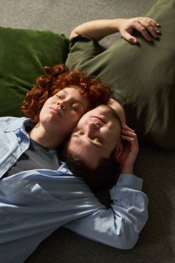 Dikey çekimde, genç çift yere uzanmış, başları birbirine değmiş, gözleri kapalı, yeşil yastıkta rahatlarken, huzurlu yüz ifadeleri sergilerken.