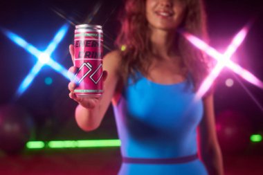 Genç beyaz kadın gülümsüyor ve elinde enerji içeceği var. Kameraya doğru canlı bir şekilde duruyor. Arka planda renkli ışıklar var.