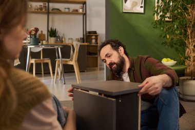 Kafkasyalı orta yaşlı bir adam mobilya montajı yaparken, parçaları hizalamaya odaklanırken, Kafkasyalı genç bir kadın modern iç mekanda gözlem yapıyor.