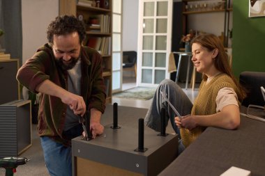 Kafkasyalı orta yaşlı bir adam tornavida kullanarak mobilya toplarken Kafkasyalı genç bir kadın elinde kullanma kılavuzuyla modern iç mekanlarda gülümsüyor.