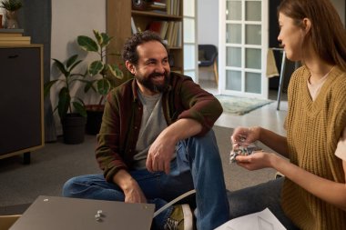 Orta yaşlı, beyaz bir adam, genç beyaz bir kadına gülümsüyor ve modern oturma odasında mobilyaları birleştiriyor.