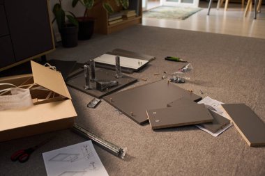 Düz paket mobilya parçaları halının üzerinde kullanma kılavuzu, vidalar, araçlar ve ambalajlar etrafa saçılmış halde duruyor, modern iç mekanda montaj süreci devam ediyor