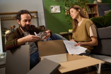 Kafkasyalı orta yaşlı bir adam, elinde donanımla yerde otururken mobilya topluyor ve yanında elinde talimat kâğıdı tutan beyaz bir kadın var.