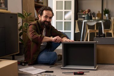 Kafkasyalı orta yaşlı bir adam mobilyaları yere diziyor, tornavida kullanıyor ve talimatları uyguluyor, aletler ve parçalarla çevrili, gülümsüyor ve modern ev içi kameralara bakıyor.