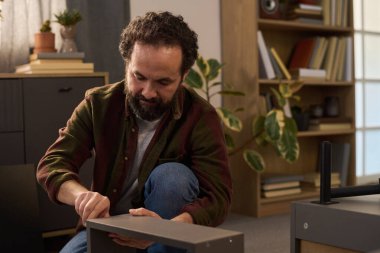 Orta yaşlı, beyaz bir adam mobilya montajı yapıyor, yerde diz çöküyor ve tornavida kullanıyor, işine odaklanmış, ev ortamında kitaplıklar ve kapalı bitkilerle çevrili.