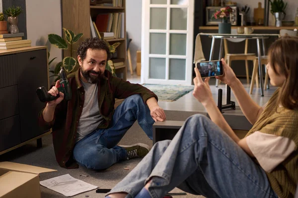 Beyaz, orta yaşlı bir adam yerde oturmuş elinde elektrikli matkapla mobilya toplarken beyaz bir kadın akıllı telefonuyla fotoğraf çekiyor.