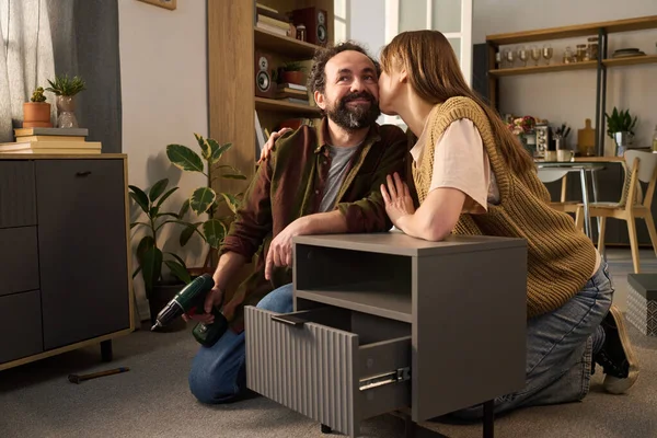 Kafkasyalı orta yaşlı bir adam, modern iç mekanlarda mobilyaların üzerine çömelmiş elektrikli matkapla mobilyaları birleştiriyor ve beyaz bir kadının yanağından öpüyor.