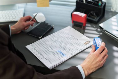 Bilinmeyen bir kadın vize başvuru belgelerini masasında işliyor, elinde onaylanmış bir pul tutuyor, vize merkezindeki modern ofis ortamında evrakları inceliyor.