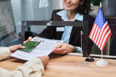 Vize merkezinde çalışan orta yaşlı beyaz bir kadın elleri görünen genç bir yetişkinden belge ve pasaport alıyor. Masada duran Amerikan bayrağı, başvuru formları.