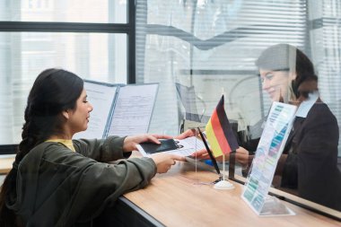 Orta yaşlı beyaz kadına pasaport ve belgeler gönderen genç Latin bir kadın vize merkezindeki camın arkasında, hem evrak işleriyle hem de masasında görünen Alman bayrağıyla etkileşimde.