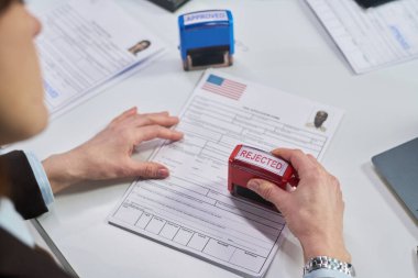 Kafkasyalı orta yaşlı bir kadın vize başvuru formunu reddedilmiş olarak damgalıyor. Vize merkezinde el ele tutuşmalar, resmi evraklar ve onay damgası.