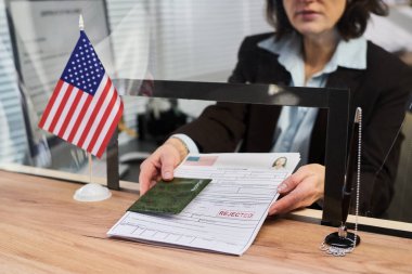 Orta yaşlı, beyaz bir kadın vize başvurusu yapıyor. Elinde reddedilmiş pul ve pasaport, masasında Amerikan bayrağı var. Vize merkezinde çalışıyor.