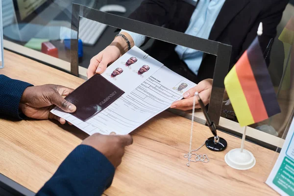 Beyaz bir kadın, vize merkezinde siyahi bir adama vize başvurusunda bulunuyor. Elleriyle pasaport ve resmi belgeleri Alman bayrağıyla değiş tokuş ediyor.