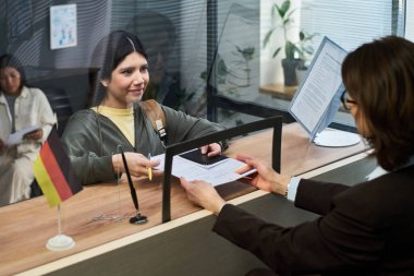 Latin genç bir kadın, orta yaşlı beyaz bir kadına vize merkezinde evrak dağıtıyor. İkisi de cam bölme, masa başında Alman bayrağı, görünür evraklar.