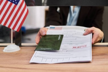 Pasaport ve vize başvuru formunu, vize resepsiyonunda reddedilmiş pulla teslim eden orta yaşlı beyaz bir kadın. Masasında Amerikan bayrağı, el ve belgelerin kapatılması.