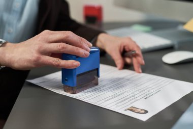 Vize merkezinde vize başvurusu belgelerini damgalayan, elinde kalem tutan kimliği belirsiz orta yaşlı bir kadın kimlik fotoğrafıyla el işleme evraklarını masasında kapatıyor.