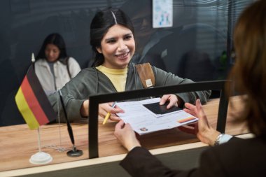 Pasaport ve vize başvuru formunu vize merkezinde tezgahın arkasında Alman bayrağı taşıyan beyaz bir kadına verirken gülümseyen genç İspanyol kadın.