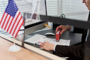 Orta yaşlı, beyaz bir kadın vize başvurusunda bulunuyor. Resmi evrakları onaylı damgalıyor. Vize merkezinde çalışıyor.