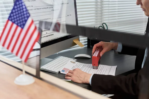 Orta yaşlı, beyaz bir kadın vize başvurusunda bulunuyor. Resmi evrakları onaylı damgalıyor. Vize merkezinde çalışıyor.
