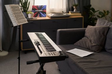 Elektronik klavye oturma odasının yanında duruyor. Masada notalar, yerde kağıtlar arka planda plak çalar ve dekoratif bitkiler var. Görünürde kimse yok.