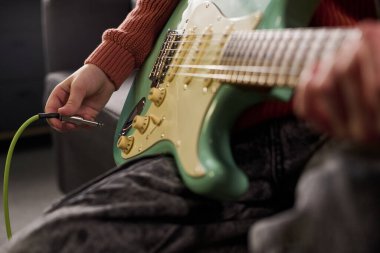 Kafkasyalı genç elektro gitar kablosunu enstrümana takıyor, evde oturup gitar ve ellere odaklanıyor ve müzik çalmaya hazırlanıyor.