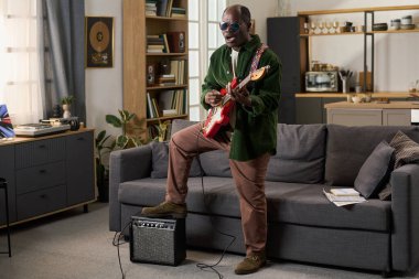 Orta yaşlı siyah adam oturma odasında dikilirken elektro gitar çalıyor, güneş gözlüğü takıyor ve şarkı söylüyor, yerde yükseltici, kitaplık ve mutfak arka planda görünüyor.