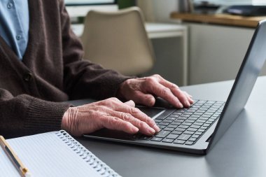 Bilgisayar okuma yazma kursu sırasında dizüstü bilgisayarda yazan, elleri klavyede görünen, masadaki spiral defter ve kalemle dijital beceri eğitimine katılan kimliği belirsiz yaşlı bir kadın.