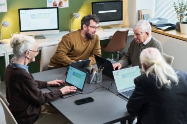 Orta yaşlı beyaz bir adam sınıftaki bilgisayar okuma yazma kursunda dizüstü bilgisayar kullanan Kafkasyalı üst düzey erkek ve kadınları eğitiyor.