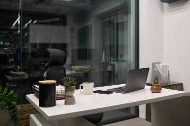 Boş ergonomik sandalyenin olduğu modern ofis çalışma alanı açık dizüstü bilgisayar, kahve fincanı, akıllı telefon, saksı bitkileri, doküman yığını, cam duvar arka planı
