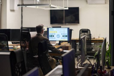 Orta yaşlı beyaz bir adam masa başında oturmuş bilgisayar üzerinde çalışıyor iş analizleri modern ofis ortamında görüntüleniyor, arka planda boş çalışma istasyonları var.
