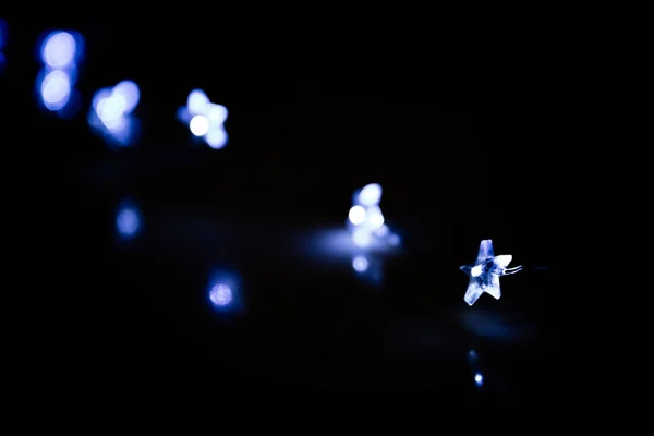 A blue electric garland glows in the dark with blue light on a black background. Blurred abstract card with one clear star-shaped light bulb. Good New Year spirit. Merry Christmas greeting card.