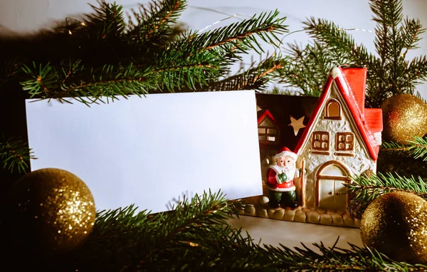 クリスマスツリーの枝 セラミックサンタクロースのおもちゃの家と光沢のあるボールと空白の名刺 新年のお祝いのコンセプトバナー ギフト券 サイドビュー イベントやパーティーへの招待 — ストック写真