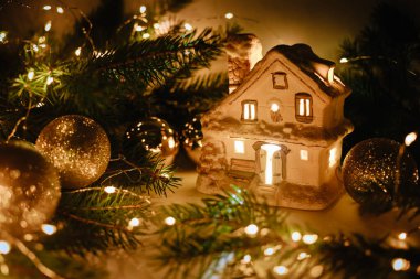Beyaz seramik ev, parlak toplar ve altın çelenk desenli Noel ağacı dalları. Sihirli duvar kağıdı. Noel şamdanı, yeni yıl 2021. Uzayı kopyala, yan görünüm. Samimi bir dekor pazarı. Oyuncak fuarı.