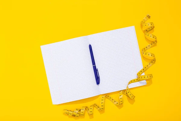 Twisted centimeter tape and clean note book and blue pen with copy space on yellow background. Greeting postcard. Body measurement. Nutrition and fitness workout plan. International Women Day 8 March.