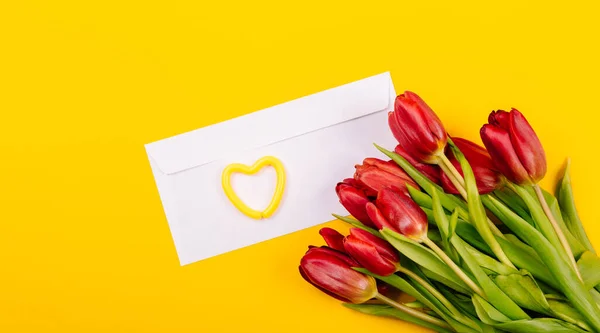 White gift certificate envelope with heart on yellow background with a bouquet of red tulips and a free place for text. Sample business or invitation card. Happy Valentine Day February 14. Woman Day.